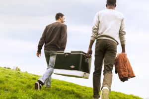Two men walk up a grassy hill on Treasure Island carrying a vintage cooler and jacket, enjoying an outdoor picnic with San Francisco Bay views.