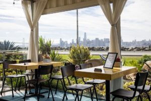 Indoor café seating on Treasure Island with large picture windows framing panoramic views of the San Francisco skyline and Bay Bridge.
