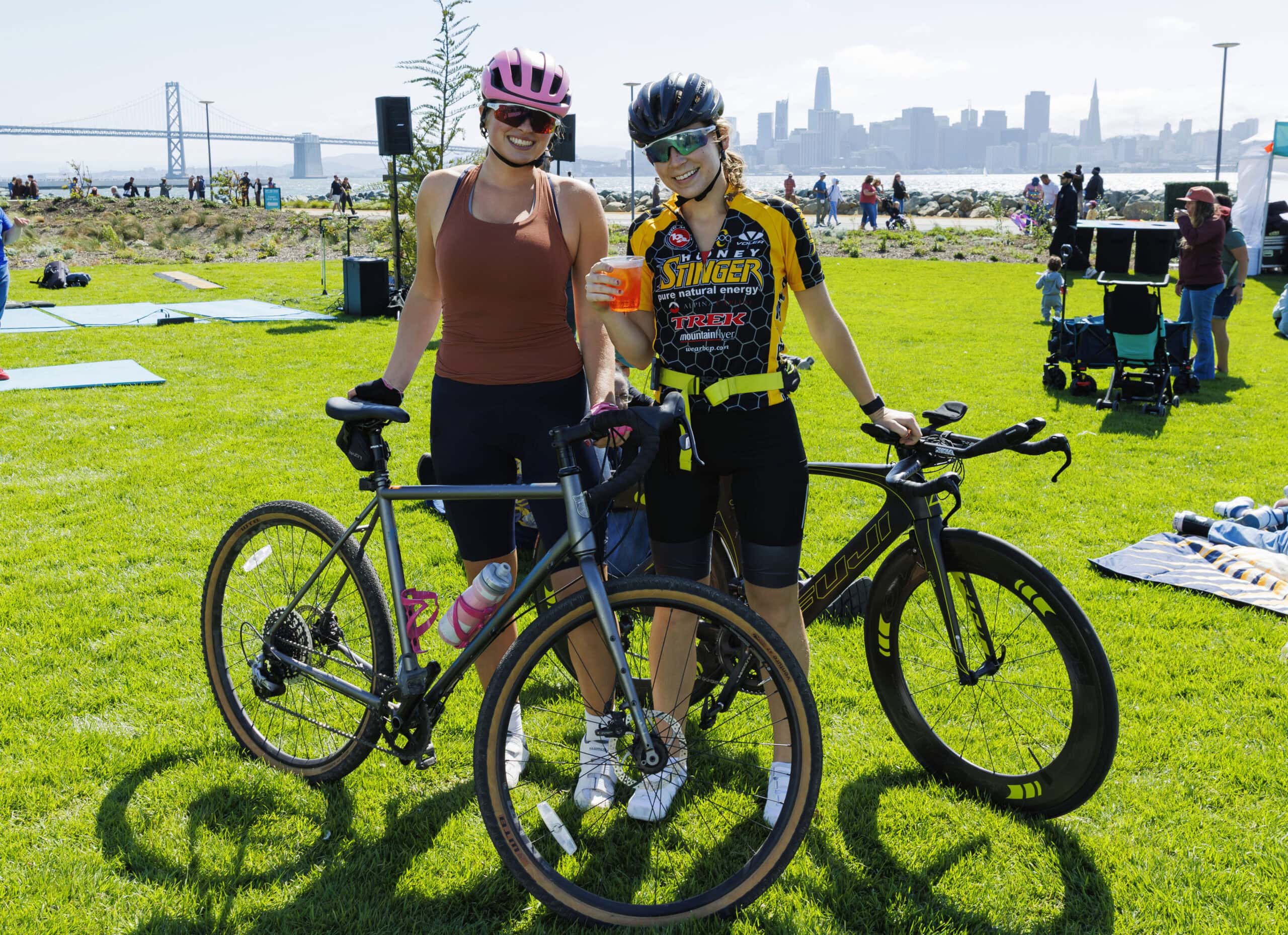 Two cyclists with road bikes smiling at a Treasure Island waterfront event with the Bay Bridge and San Francisco skyline in the background.