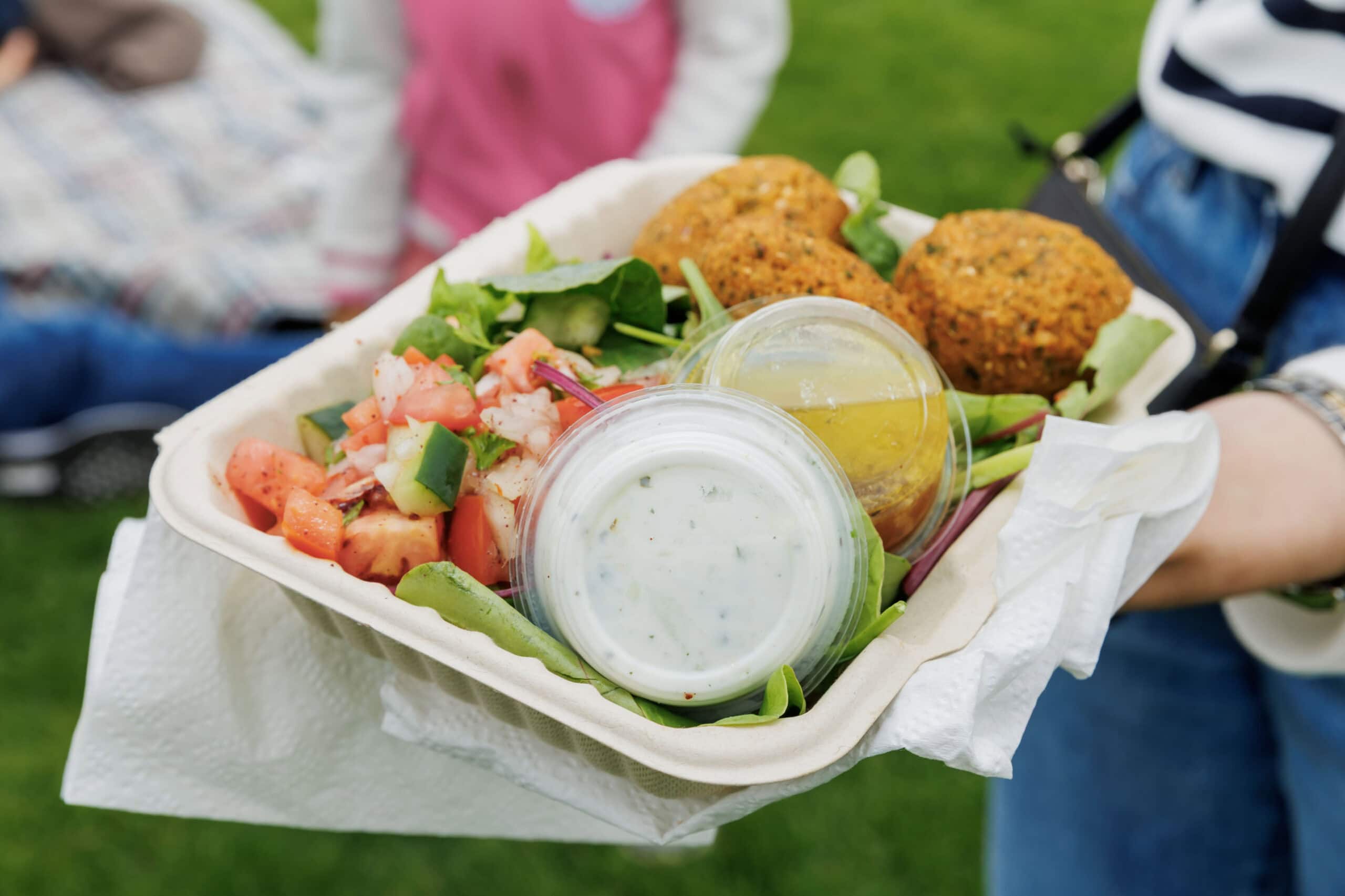 Fresh salad with falafel, mixed greens, diced tomatoes, cucumbers, and dressing served in a compostable tray at an outdoor waterfront event.
