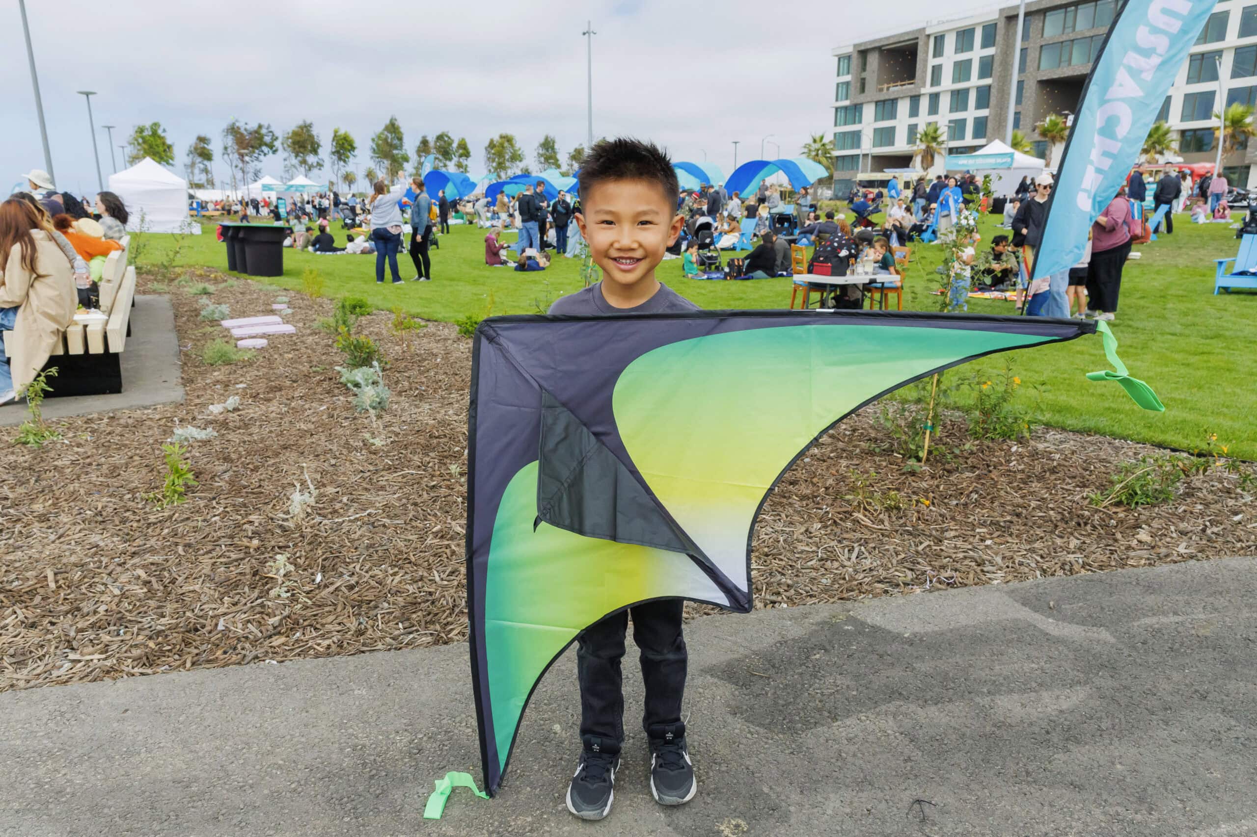 Smiling child holding a green kite at a Treasure Island waterfront park event with families, tents, and modern apartments in the background.