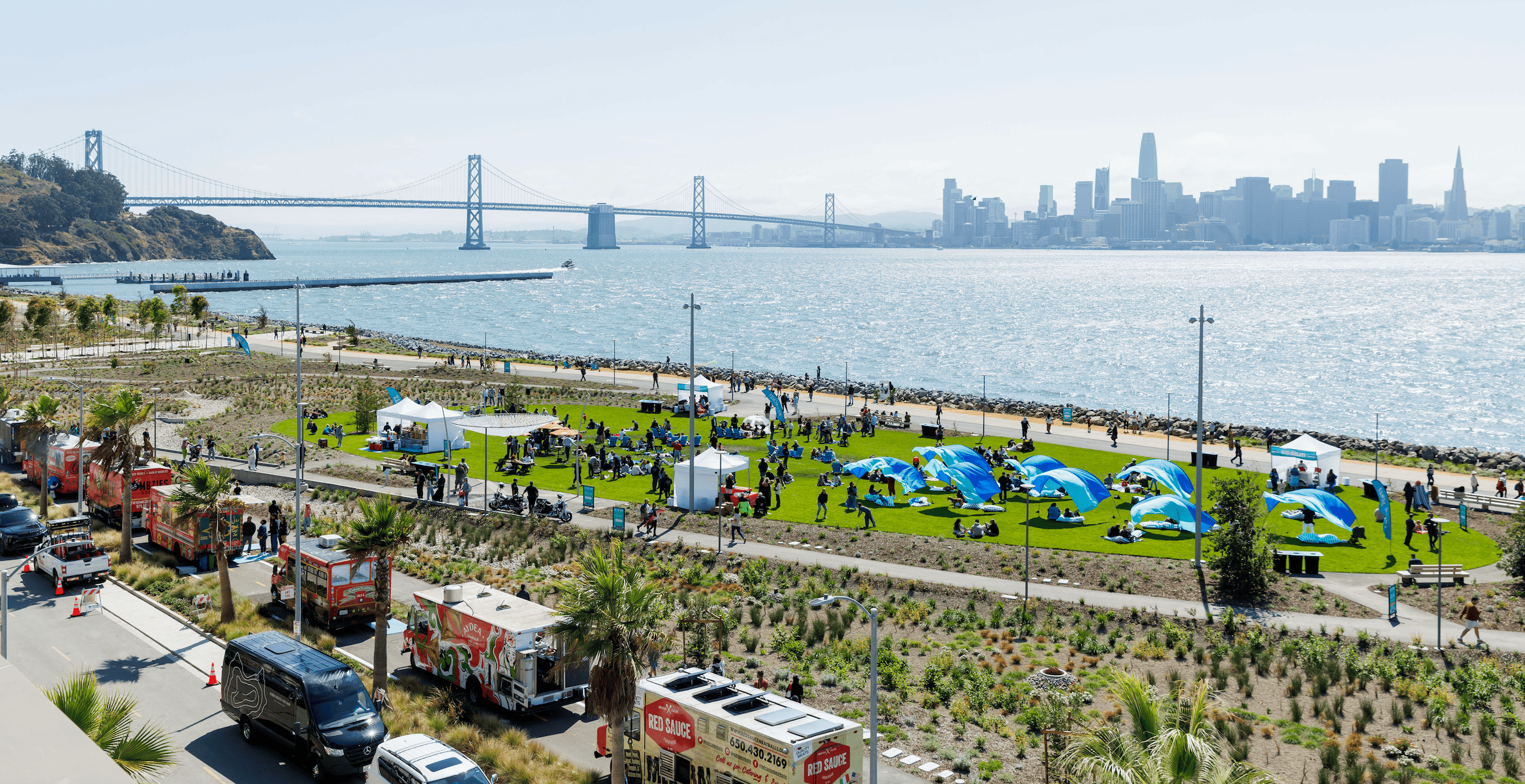 Aerial view of a Treasure Island waterfront park event with food trucks, blue shade tents, the Bay Bridge, and the San Francisco skyline in the background.