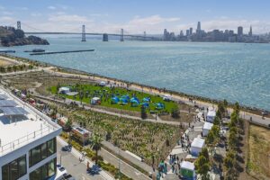 Aerial view of Cityside Park grand opening on Treasure Island with event tents, blue shade structures, the Bay Bridge, and the San Francisco skyline in the background.