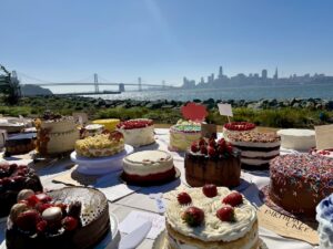 Assorted decorated cakes on display at a waterfront dessert event on Treasure Island with the Bay Bridge and San Francisco skyline in the background.