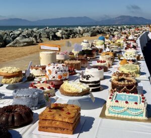 Long dessert table with dozens of decorated cakes displayed along the Treasure Island waterfront with bay and mountain views in the background.