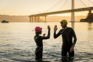 Two open water swimmers in wetsuits high-fiving in the San Francisco Bay at sunrise with the Bay Bridge in the background.
