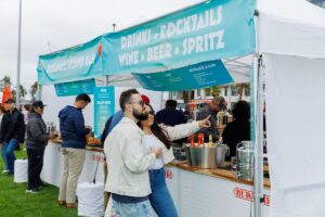 Couple ordering drinks at a Treasure Island outdoor bar tent offering cocktails, wine, beer, and spritz during a waterfront festival.