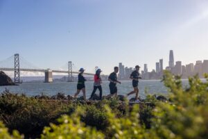 Group of runners jogging along the Treasure Island waterfront trail with the Bay Bridge and San Francisco skyline in the background.