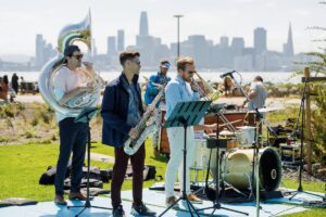 Live jazz band performing outdoors at a waterfront park with the San Francisco skyline in the background on a sunny day.