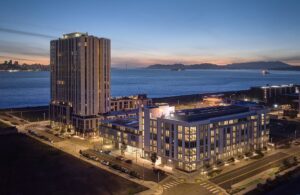 Twilight aerial view of modern waterfront apartment buildings on Treasure Island with the San Francisco Bay, Bay Bridge, and city skyline illuminated at dusk.