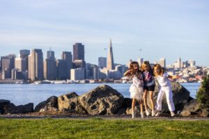 Three children jumping on a waterfront path at Treasure Island with the San Francisco skyline and Transamerica Pyramid in the background.