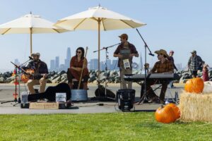 Live country band performing outdoors on a grassy lawn with pumpkins and patio umbrellas during a daytime fall festival event.