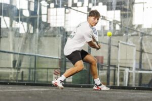Male tennis player hitting a two-handed backhand on an indoor hard court during an intense practice session.