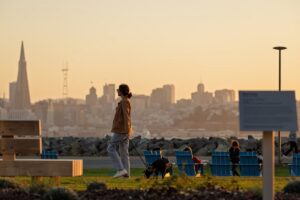 Woman walking her dog at sunset in a waterfront park with the San Francisco skyline and Transamerica Pyramid silhouetted in the background.