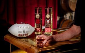 Hand holding a glass of whiskey on a wooden bar next to a San Francisco 49ers football and premium liquor bottles in the background.