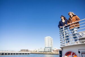Couple standing on a boat deck railing overlooking a city waterfront skyline under a clear blue sky.