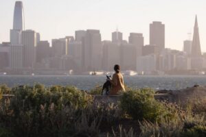 Person sitting on a grassy overlook with a dog, facing the San Francisco skyline and Transamerica Pyramid across the bay at sunset.