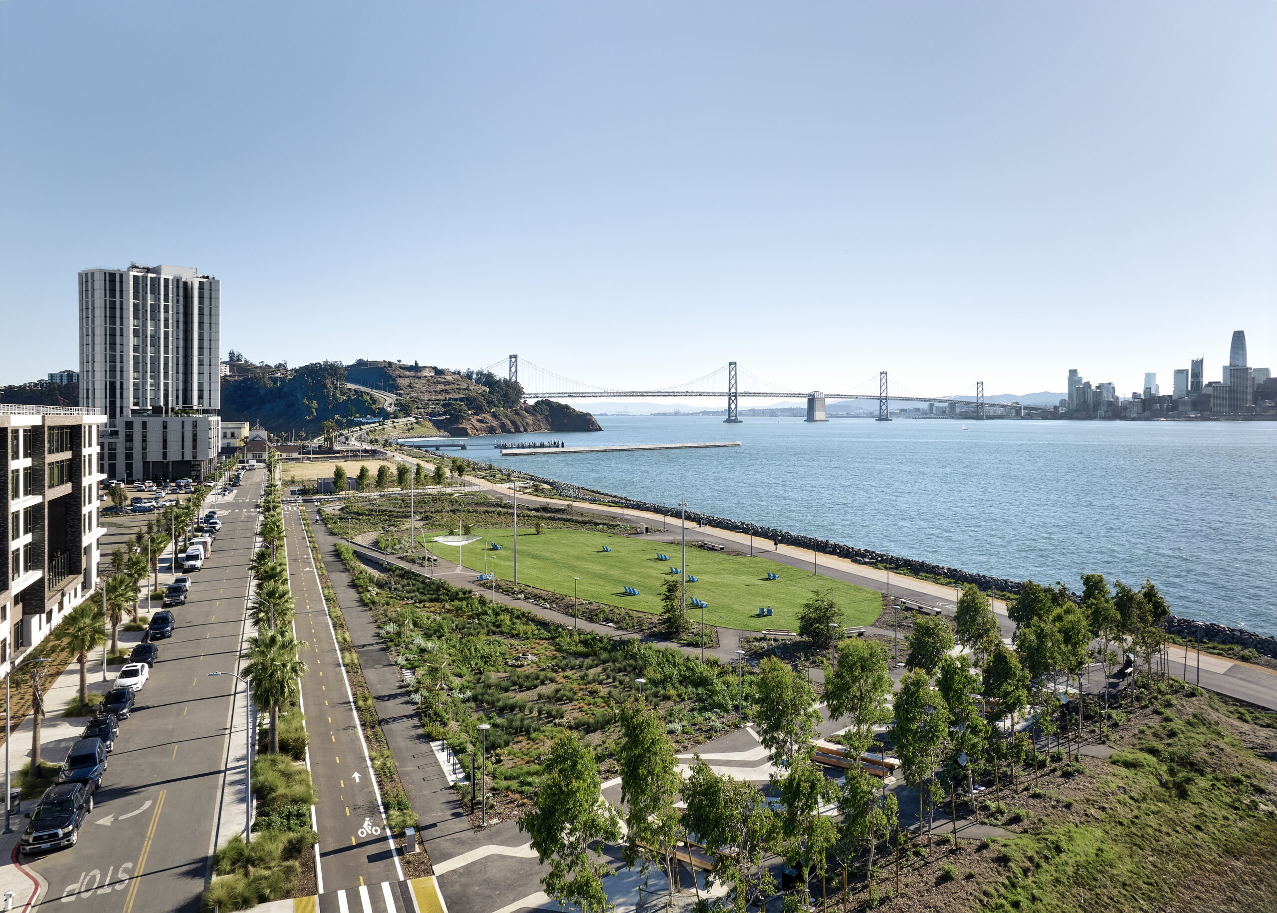 A wide aerial view of a waterfront park along a bay, with a landscaped green space, walking paths, and a bike lane running beside a road lined with palm trees. In the distance, a long suspension bridge spans the water toward a city skyline with modern high-rise buildings under a clear blue sky.