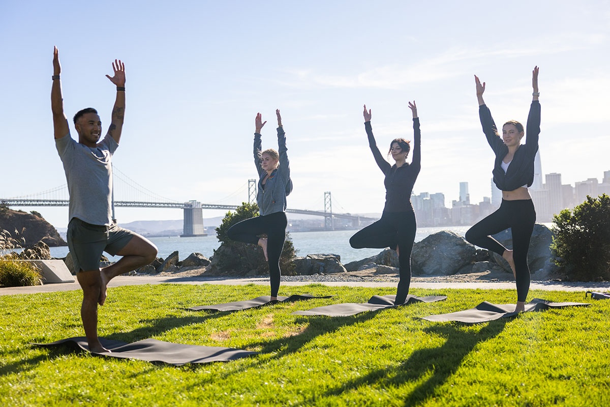 A small group practicing yoga on mats in a grassy park beside the water, with a bridge and city skyline in the background.