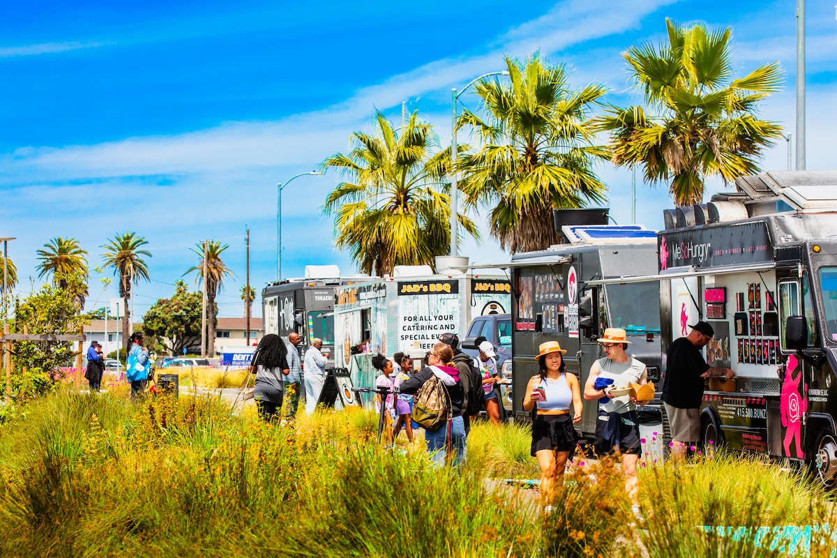 People walking and gathering around colorful food trucks lined along a sunny street with palm trees and greenery.