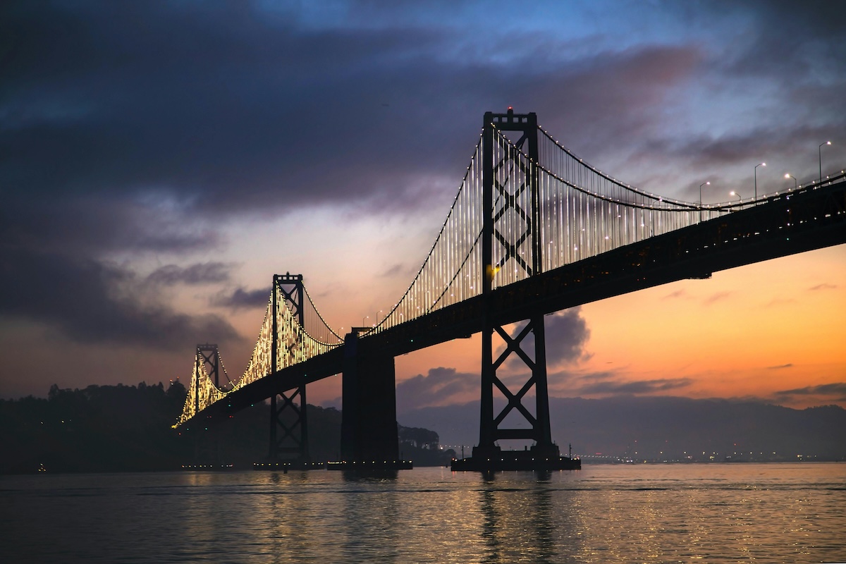 A suspension bridge lit with a string of lights spans across a body of water at sunset, with a warm orange glow on the horizon and dark silhouettes of the bridge towers against a cloudy sky.