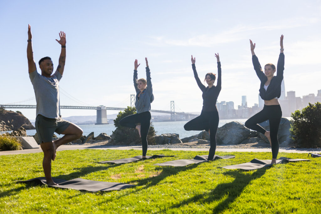 People practicing yoga on mats by the waterfront, with the Bay Bridge and San Francisco skyline in the background.
