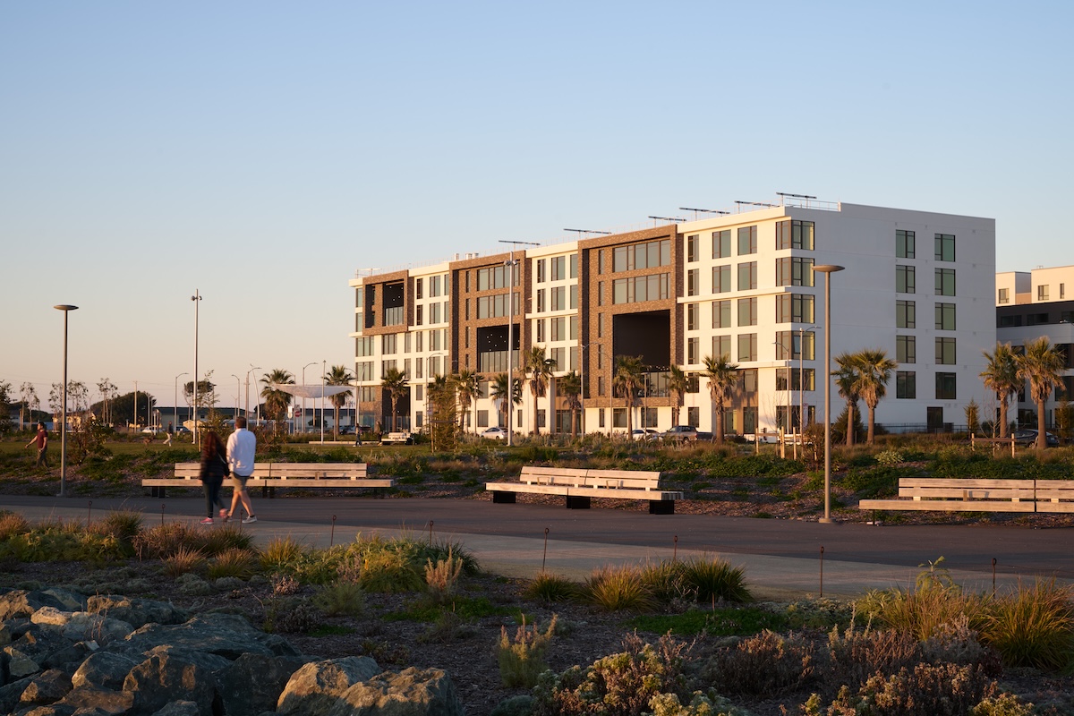 Contemporary mid-rise residential building bordered by palm trees, walking paths, landscaped gardens, and a waterfront promenade under clear skies.