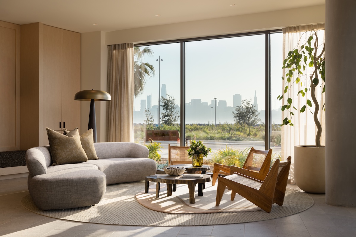 Bright, modern living room with floor-to-ceiling windows framing views of the waterfront park and the San Francisco skyline, featuring contemporary furnishings and abundant natural light.