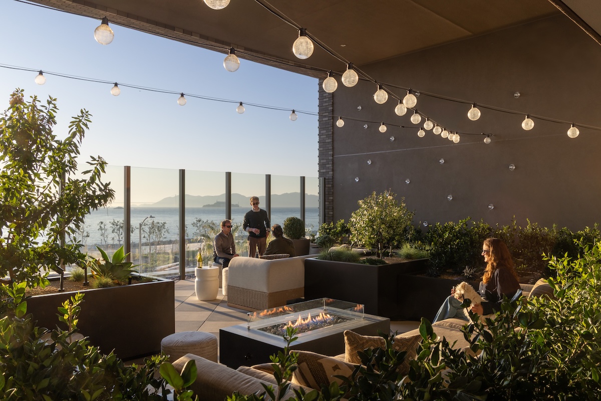 Rooftop lounge with a fire pit, string lights, lush planters, and comfortable seating, where residents gather while enjoying bay and skyline views.