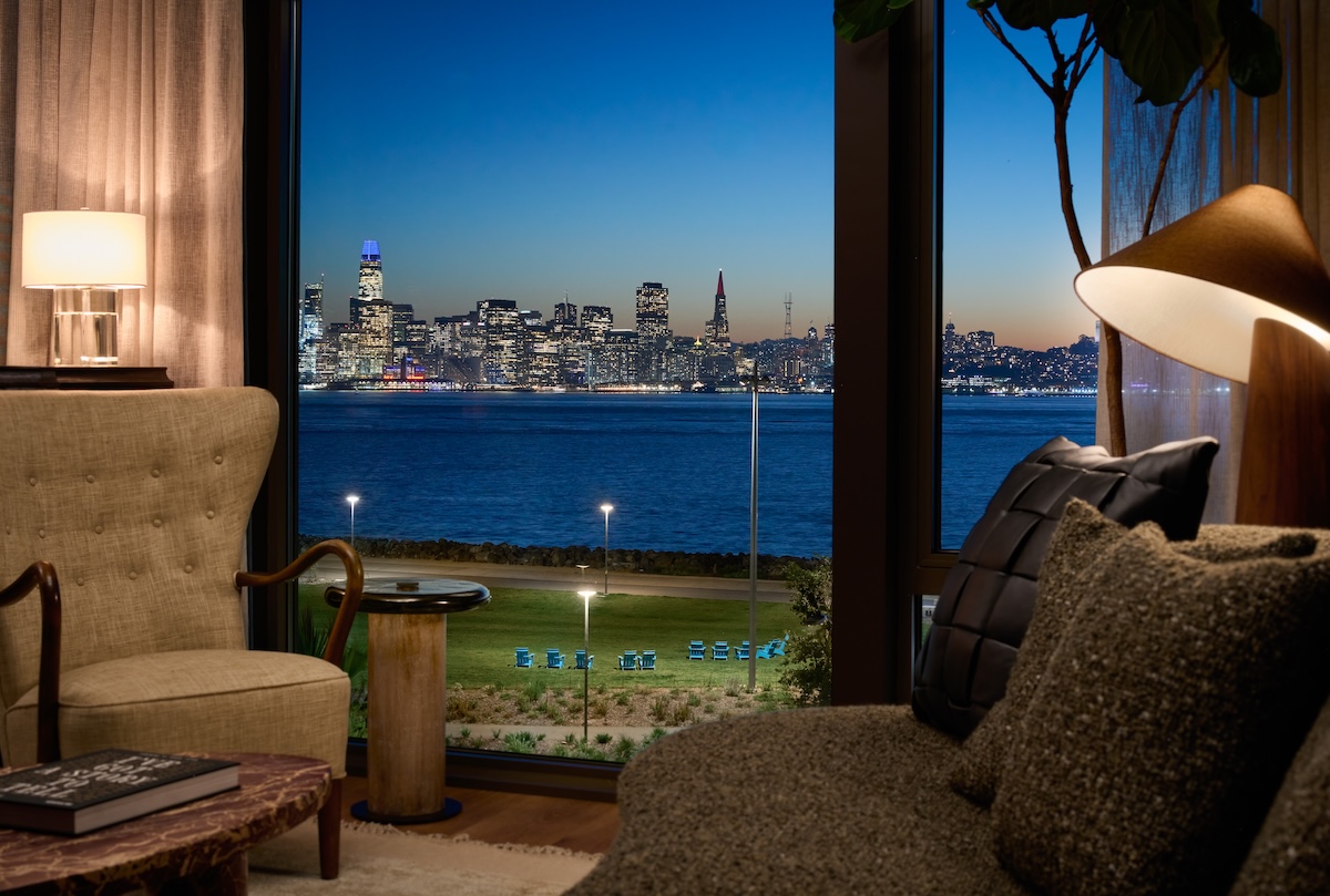 Cozy living room with large picture windows showcasing illuminated views of the San Francisco skyline across the bay at night, furnished with comfortable seating and warm ambient lighting.