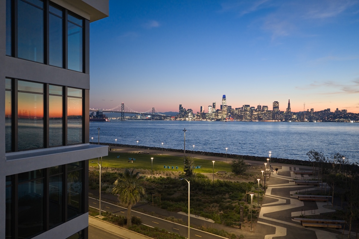 Modern waterfront apartment building overlooking a landscaped park and the San Francisco skyline across the bay at dusk, with the Bay Bridge visible in the distance.